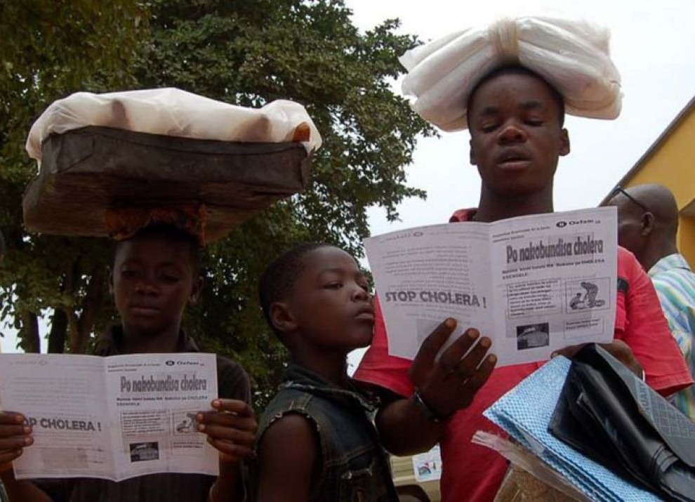 Children reading cholera-prevention leaflets warning about the dangers of cholera and how it can be prevented.